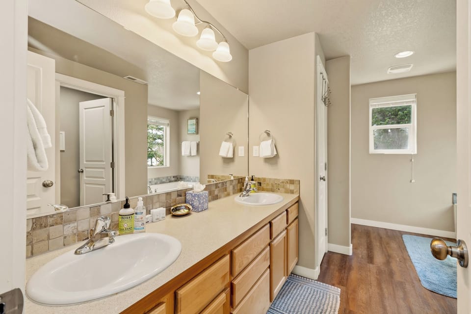 Your spacious bathroom features warm wood cabinetry, a stylish tiled backsplash, and a large mirror reflecting abundant natural light from the private water closet window.