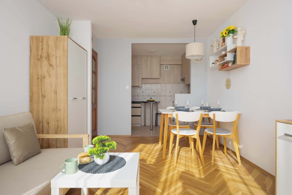 The living area is shown looking toward the dining nook and kitchenette, emphasizing the open and airy layout. Sunlight streams through large windows, highlighting the light wood furniture and herringbone flooring.