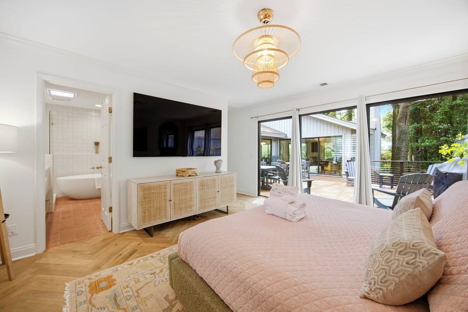 Golden oak floors and rattan textures glow under a sculptural pendant lamp in this airy bedroom. Sliding glass doors open directly to the deck, while a soaking tub awaits just steps away across the terracotta tile.