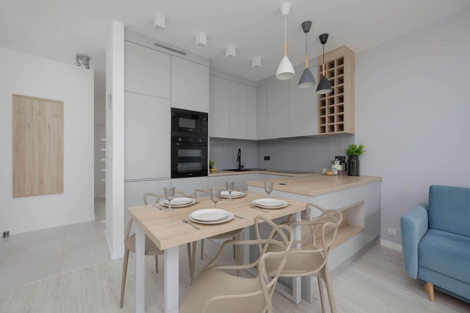 A bright dining area with a wooden table set near the kitchenette and living room. Neutral colors and modern furnishings create a cozy interior.