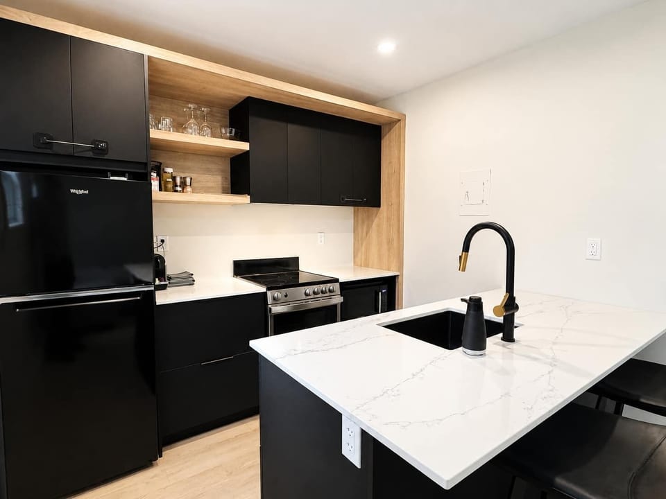 Une cuisine contemporaine haut de gamme dotée d'un îlot blanc marbré avec un évier noir intégré et un robinet aux accents dorés, d'armoires noir mat épurées et d'une cuisinière moderne en acier inoxydable.