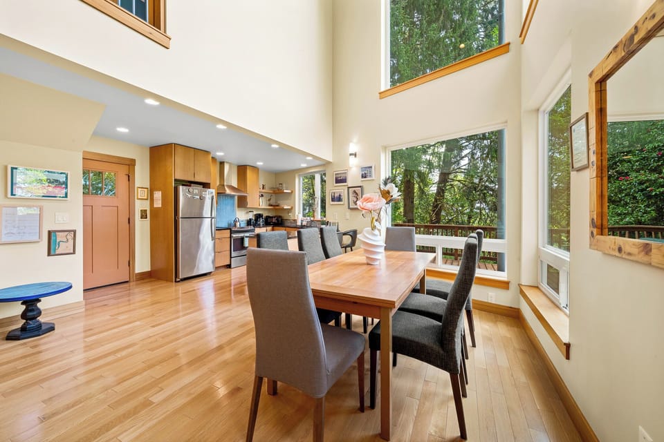 Sunlight pours through soaring windows onto warm wood floors, highlighting your dining space that opens to a fully equipped kitchen—perfect for gathering over home-cooked meals surrounded by nature.