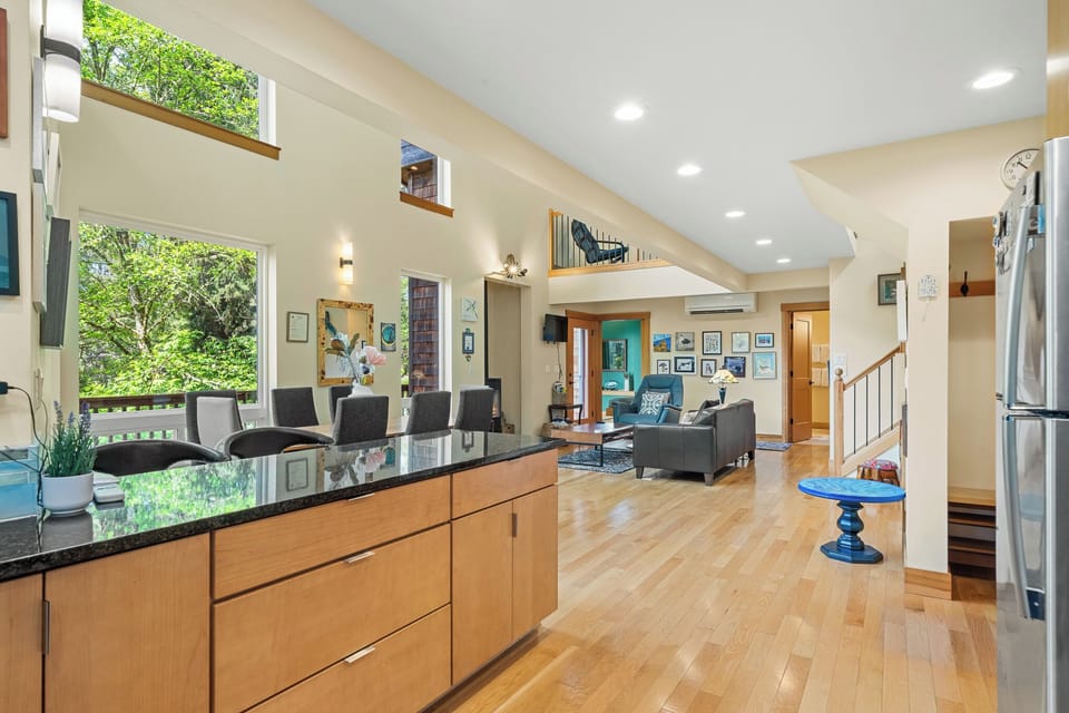 Sunlight pours through tall windows into your open-concept living space, where gleaming hardwood floors connect the modern kitchen to a cozy seating area. The vaulted ceilings and natural greenery views create an airy, welcoming retreat.