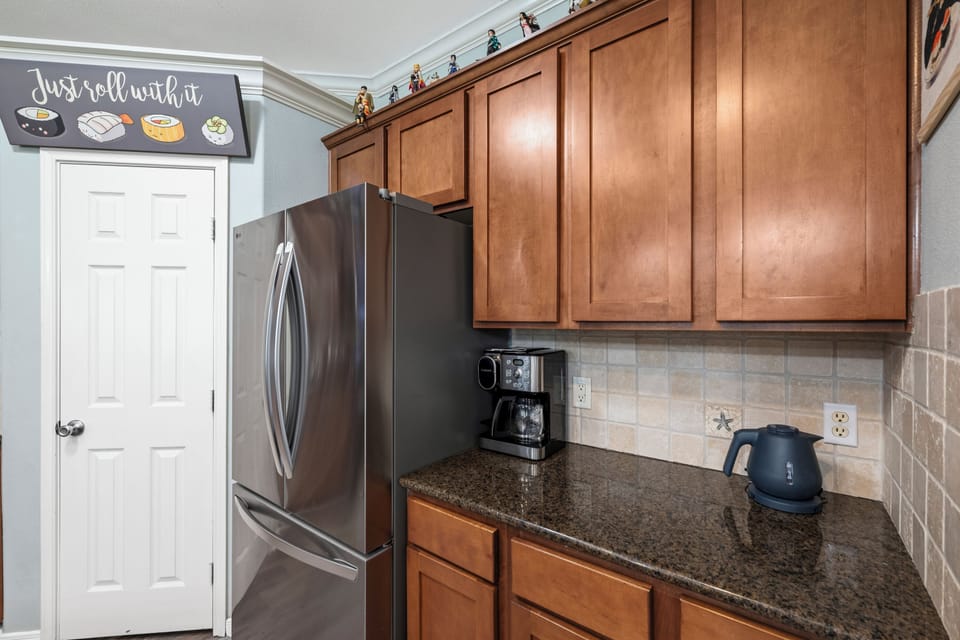 Your morning coffee station awaits with a stainless steel refrigerator, rich wood cabinetry, and modern appliances on sleek granite countertops—everything you need for a cozy start.