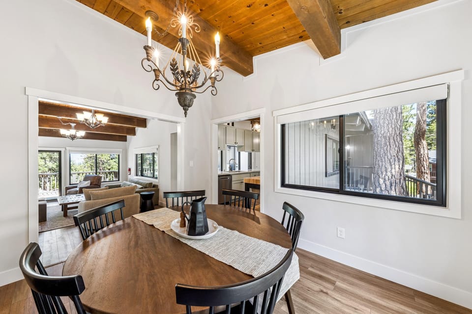 Round wood dining table with black spindle-back chairs beneath a wrought iron chandelier and exposed wood ceiling beams.