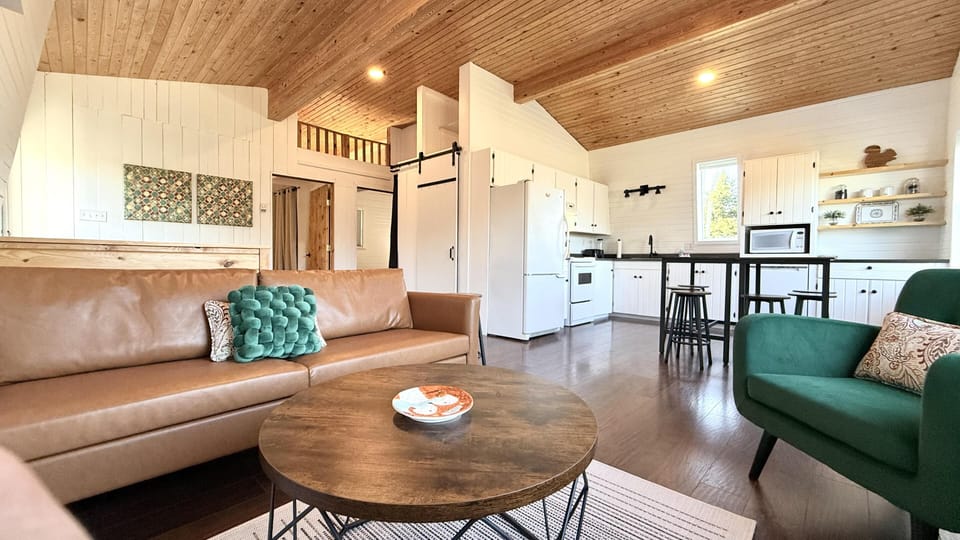 Upper level.  Open-concept living area with tan leather sofa, green armchair, round wood coffee table, and white kitchen cabinets.