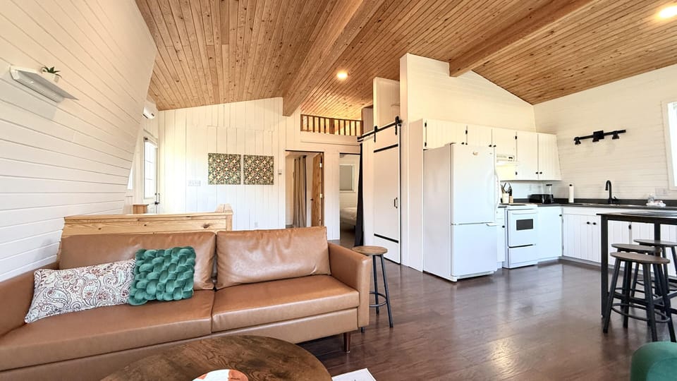 Open-concept living area with brown leather sofa, white shiplap walls, vaulted wood ceiling, and white kitchen cabinetry.