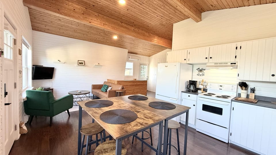Open-concept kitchen and dining area with white cabinetry, wood plank ceiling, and rustic wood table with stools.
