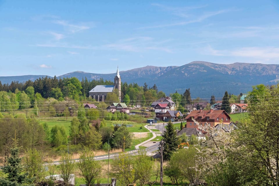 A panoramic view shows the property nestled in a picturesque valley with high mountains rising in the distance. The scene is dotted with traditional alpine-style buildings and vibrant spring foliage.