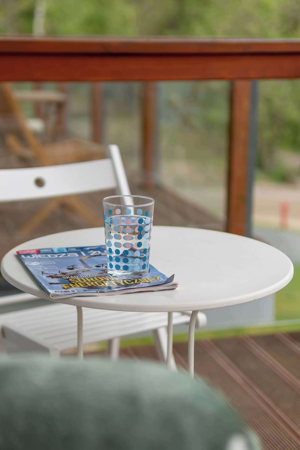 A small white table on the balcony holds a refreshing drink and a magazine for a relaxing afternoon. The glass railing ensures an unobstructed view of the greenery beyond.