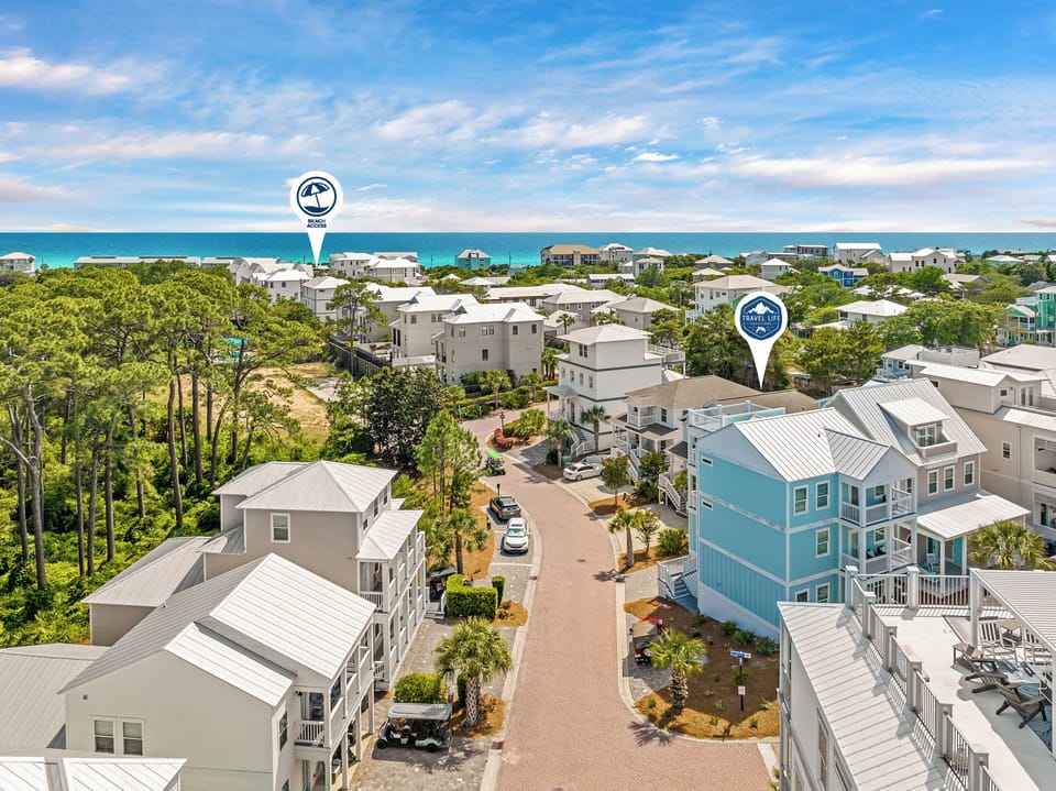 Aerial view of a coastal vacation rental community featuring modern beach homes with metal roofs, surrounded by lush greenery and just steps from turquoise waters.