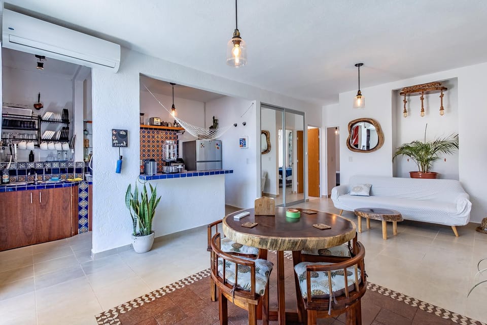 Open living and dining area with round wood table, cushioned chairs, white sofa, and blue-tiled kitchen pass-through.