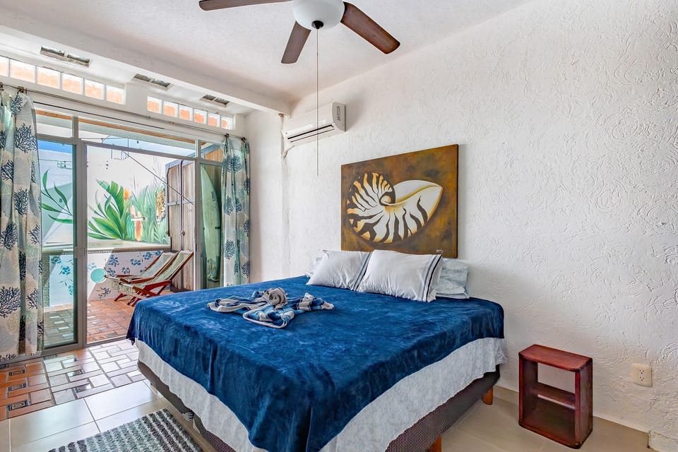 King bed with navy blanket and shell artwork, sliding glass door to balcony with wooden lounge chairs.