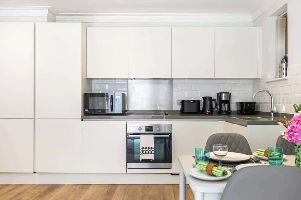 Cook in style in this modern open kitchen, featuring sleek off-white flat-panel cabinetry, white subway tile backsplash, integrated appliances including an oven and microwave, counter appliances, and a set dining table in the foreground.