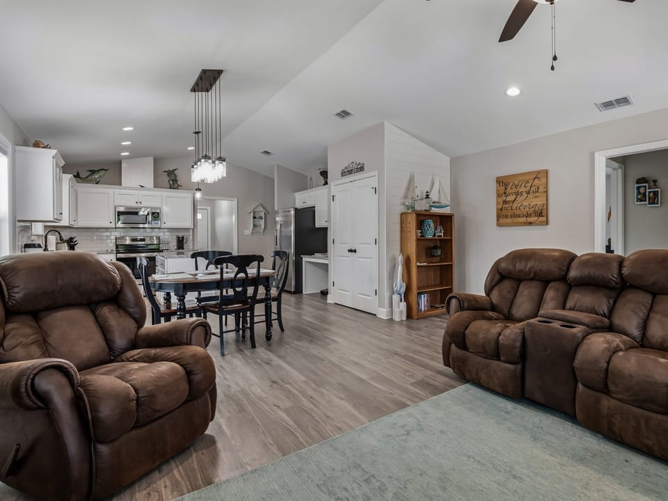 Open-plan living, dining, and kitchen with brown sofas, ceiling fan, and a bookshelf with coastal decor