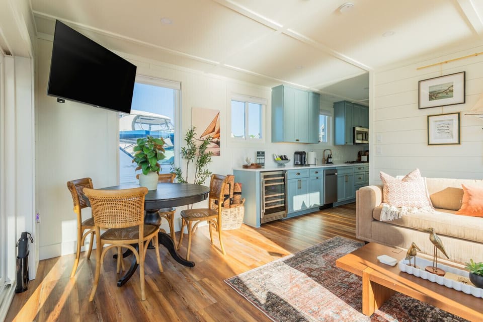 The open living and dining area, with a round dining table for four, the blue-gray galley kitchen and wine fridge beyond, and marina views filling the windows throughout.