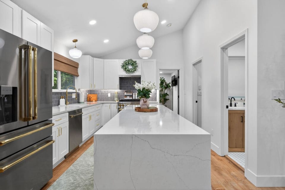Show-stopping chef`s kitchen featuring a massive waterfall quartz island, designer pendant lighting, vaulted ceilings, and stainless appliances with brass accents. White shaker cabinets and custom tile create a bright, luxurious gathering space