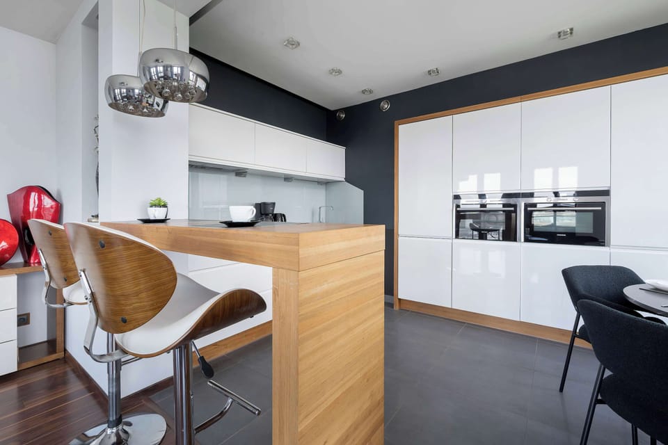 The breakfast bar area features a warm wooden countertop and modern bar stools with curved wooden backs. The kitchen in the background is equipped with high-end integrated ovens and white cabinetry.