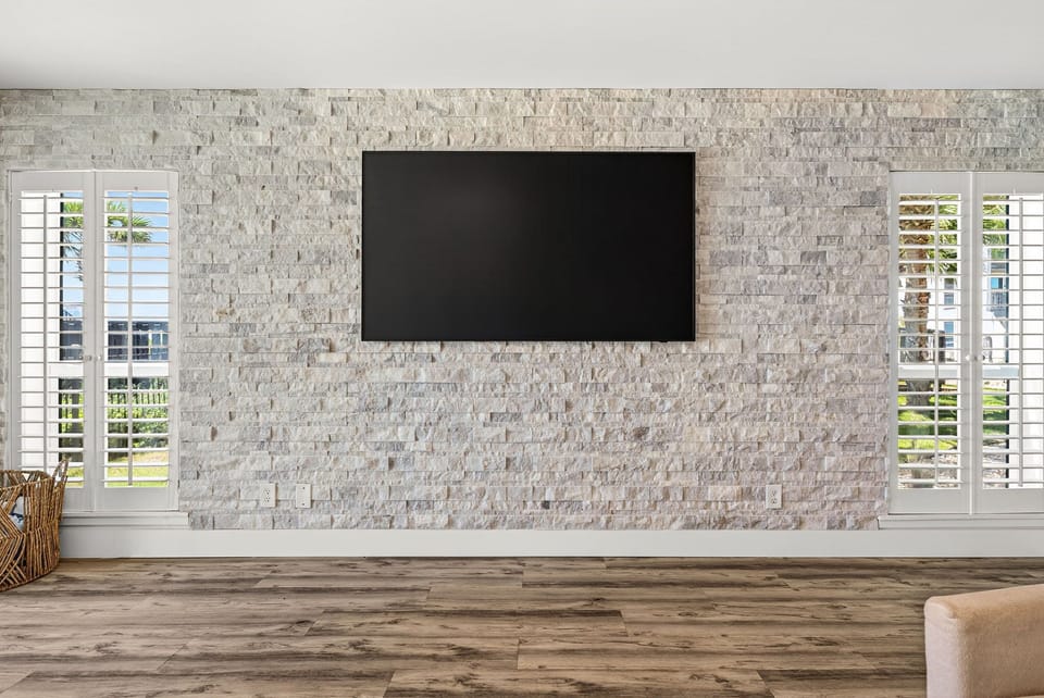 A bright open living space with textured stone accent wall and wall-mounted TV, natural light streaming through plantation shutters, and warm hardwood floors that invite you to relax.