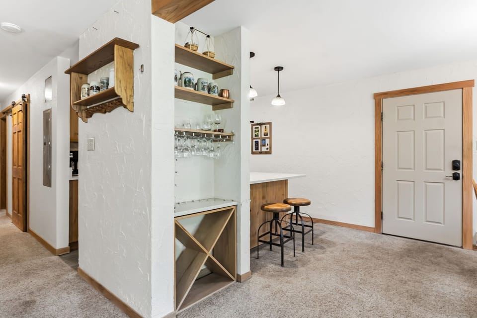 Step into a charming space where rustic wood shelves display thoughtful touches above a cozy wine nook. Warm pendant lights and industrial stools create the perfect spot for your evening unwind.