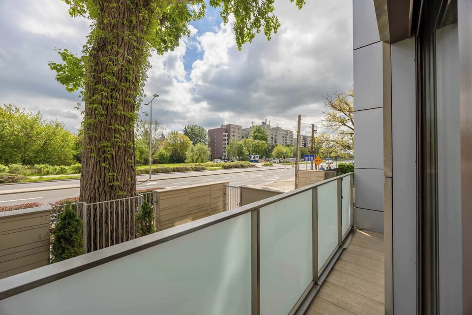 A balcony with glass railings overlooking trees and a quiet residential street. Outdoor space extends from the apartment through large sliding doors.
