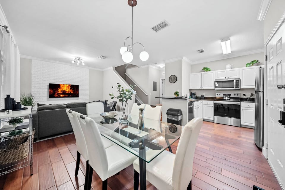 Welcome to a bright kitchen and dining area featuring glossy white cabinets and rich hardwood floors. The glass dining table with sleek chairs invites meals, while modern appliances and a cozy fireplace create a warm atmosphere.