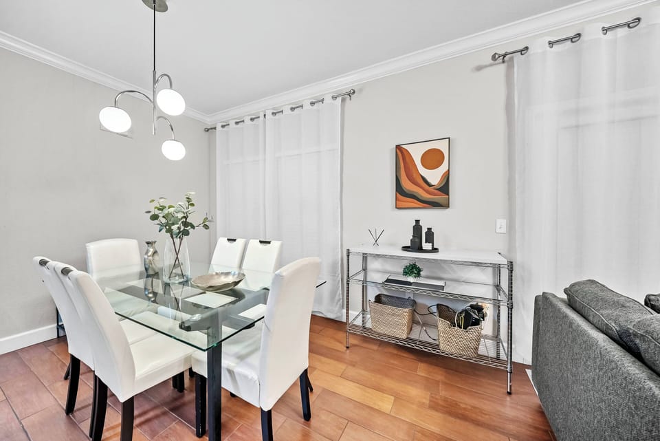 This bright dining area boasts a sleek glass table with modern white chairs, ideal for shared meals. Large windows with sheer curtains let in natural light, enhancing the welcoming atmosphere. Enjoy every moment together!