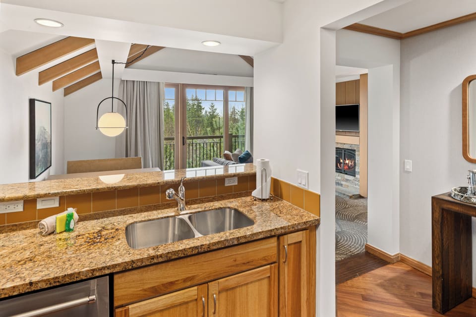 Fully equipped kitchen featuring granite counters, stainless steel sink, and warm wood cabinetry  with a cozy fireplace visible in the adjoining room.