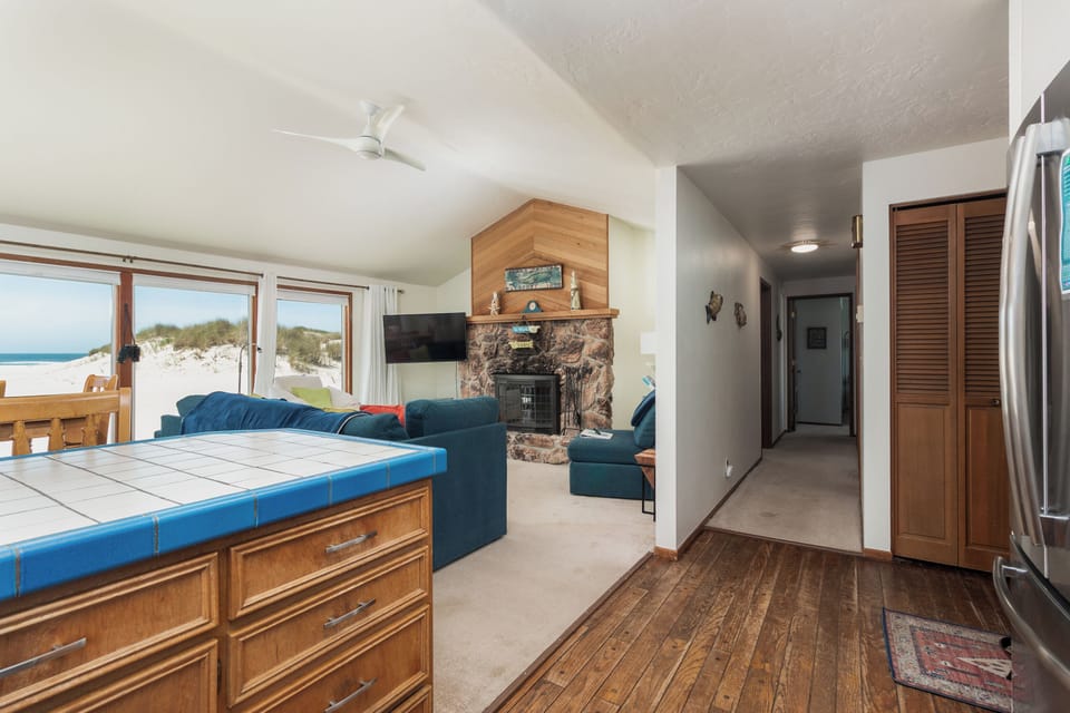 Bright coastal living space where you can unwind by the stone fireplace or watch waves roll in through expansive windows. A tiled island and cozy seating invite relaxing evenings steps from the shore.