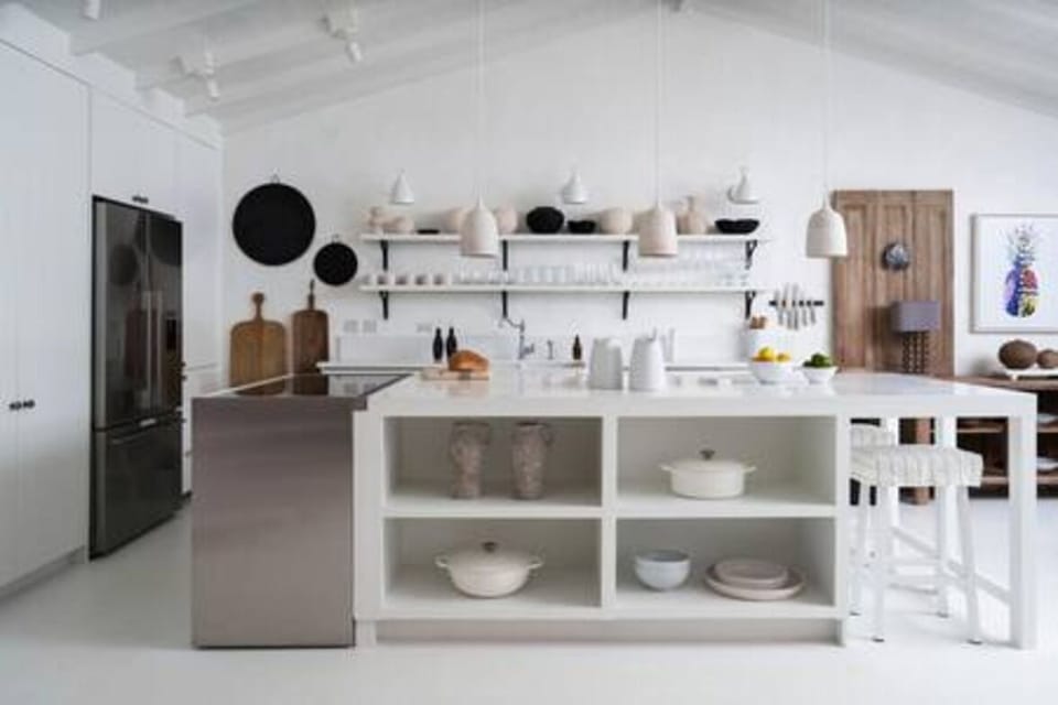 This open, airy kitchen-dining space features vaulted ceilings, a large white island with open shelving, and natural wood accents. Stylish décor, modern appliances, and sliding glass doors create a seamless connection to the lush tropical outdoors.