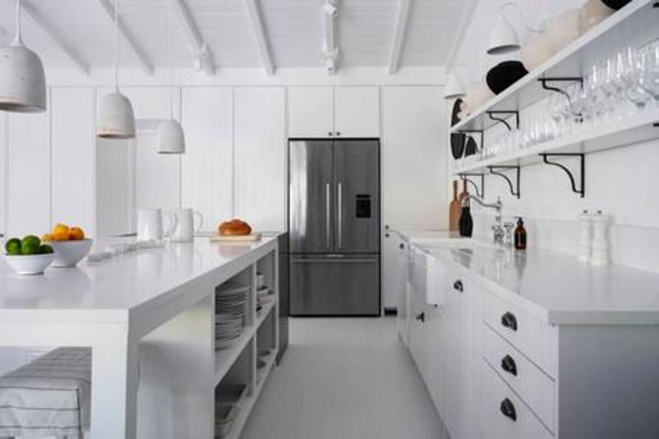 This open, airy kitchen-dining space features vaulted ceilings, a large white island with open shelving, and natural wood accents. Stylish décor, modern appliances, and sliding glass doors create a seamless connection to the lush tropical outdoors.