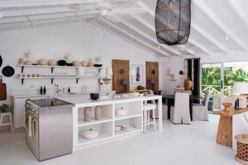 This open, airy kitchen-dining space features vaulted ceilings, a large white island with open shelving, and natural wood accents. Stylish décor, modern appliances, and sliding glass doors create a seamless connection to the lush tropical outdoors.