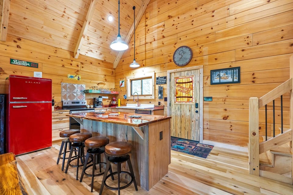 Kitchen with polished wood island, leather-topped stools, red retro fridge, and stained glass door under vaulted pine ceilings.