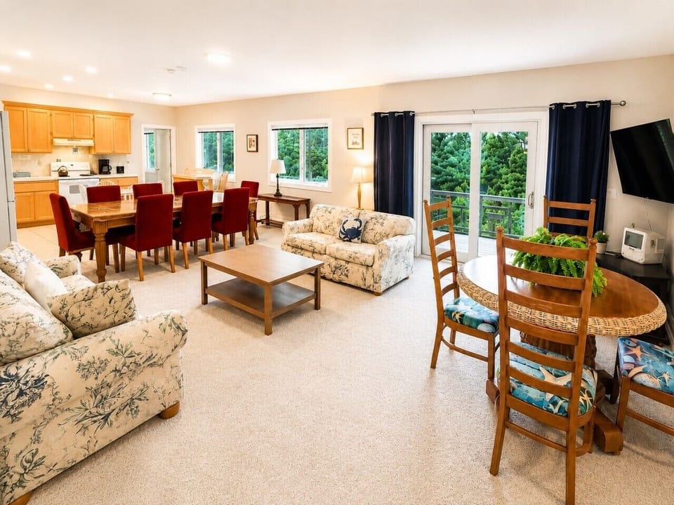 Wide-angle view of the cottage open-plan – full kitchen, large dining table with red chairs, floral sofas, and deck access.