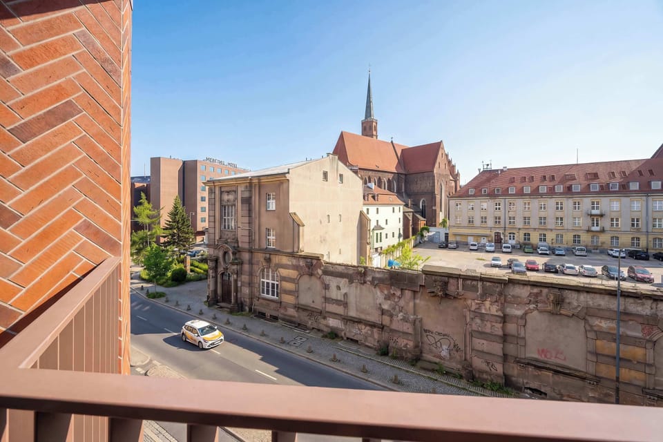 A daytime view from the balcony overlooking a city street with a magnificent historic brick building under a clear blue sky. The contrast between old architecture and modern living is clearly visible.