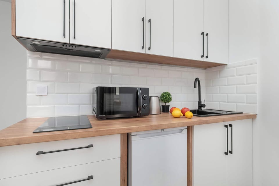 A modern kitchen corner with a microwave, kettle, and plenty of storage space. The clean white tiles and wooden shelves give it a fresh look.