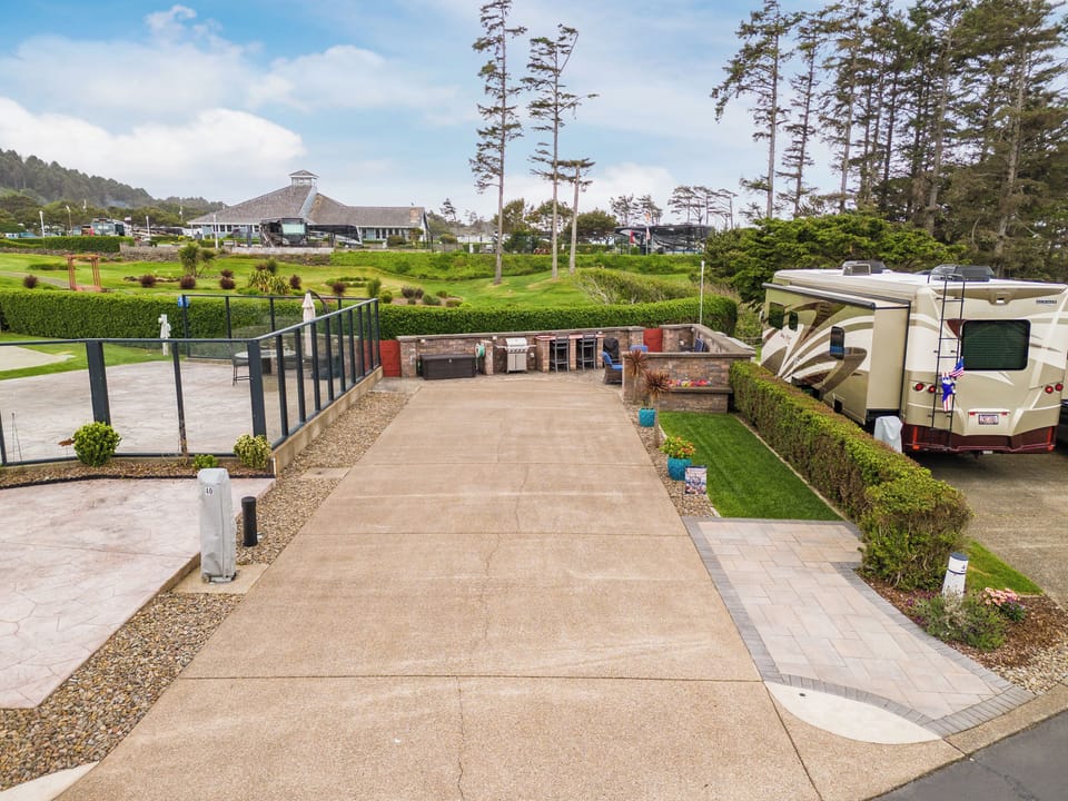 Spacious RV site with level parking, BBQ facilities, and lush landscaping surrounded by towering coastal pines and rolling green grounds.