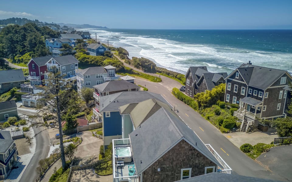 Aerial view of a coastal neighborhood with ocean waves rolling in along the shore. The property is situated steps from the beach in a scenic residential area.