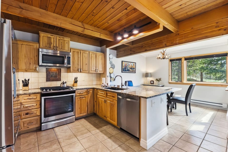 Your spacious kitchen invites you to cook and gather, featuring warm wood cabinetry, stainless steel appliances, ample counter space, and natural light streaming in through large windows overlooking greenery.