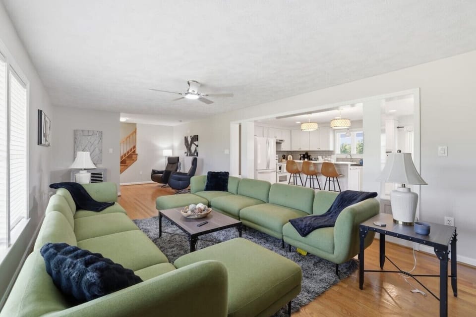 Sun‑filled living room with two plush sectionals, ceiling fan, a sleek electric fireplace, and a wall‑mounted Smart TV. Natural light and an open layout give the space a bright, elevated feel.