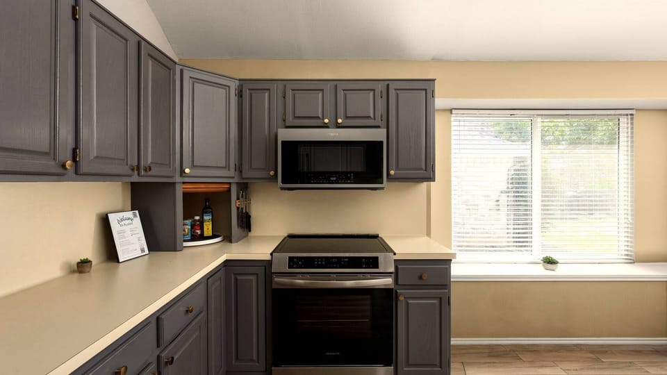 Well-appointed kitchen with bold charcoal cabinetry, gold hardware, stainless steel appliances including an electric range and over-the-range microwave, and a sunny window — a functional and stylish space ready for everyday cooking.