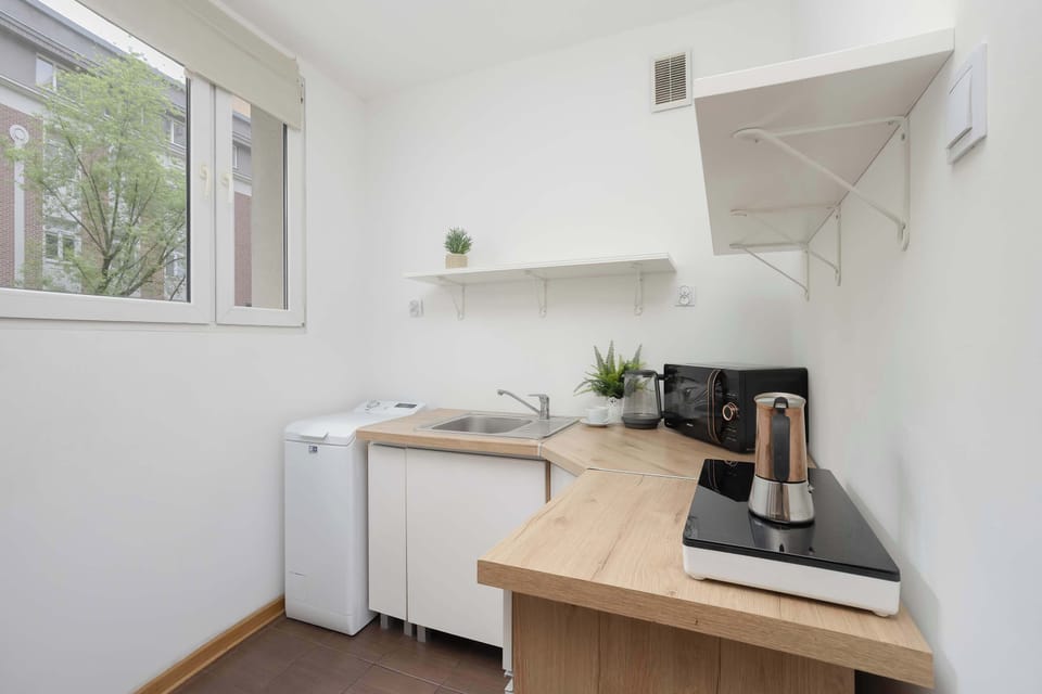 A compact kitchenette with white cabinets, wooden countertops, and a coffee machine placed beneath the window. Functional design and natural lighting make the space feel bright and practical.