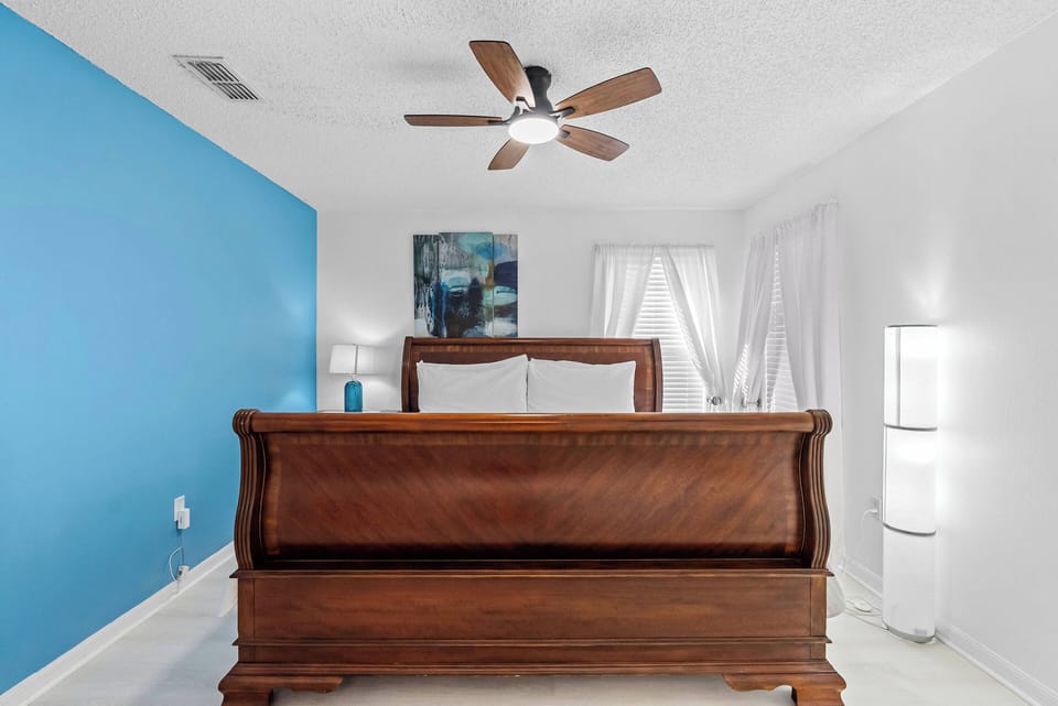 This bright bedroom features a stunning blue accent wall and a warm wooden sleigh bed. Relax in soft natural light filtered by sheer curtains, and enjoy a stylish ceiling fan for a refreshing breeze during your stay.