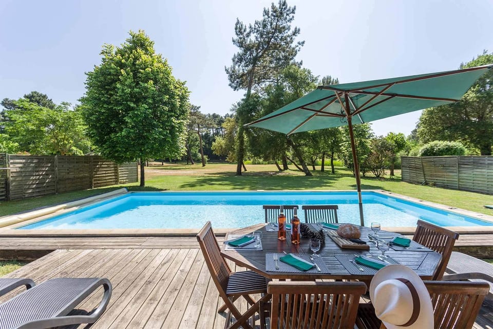 Poolside dining under a parasol on the timber deck — the garden backs onto the pines with the lake close by.