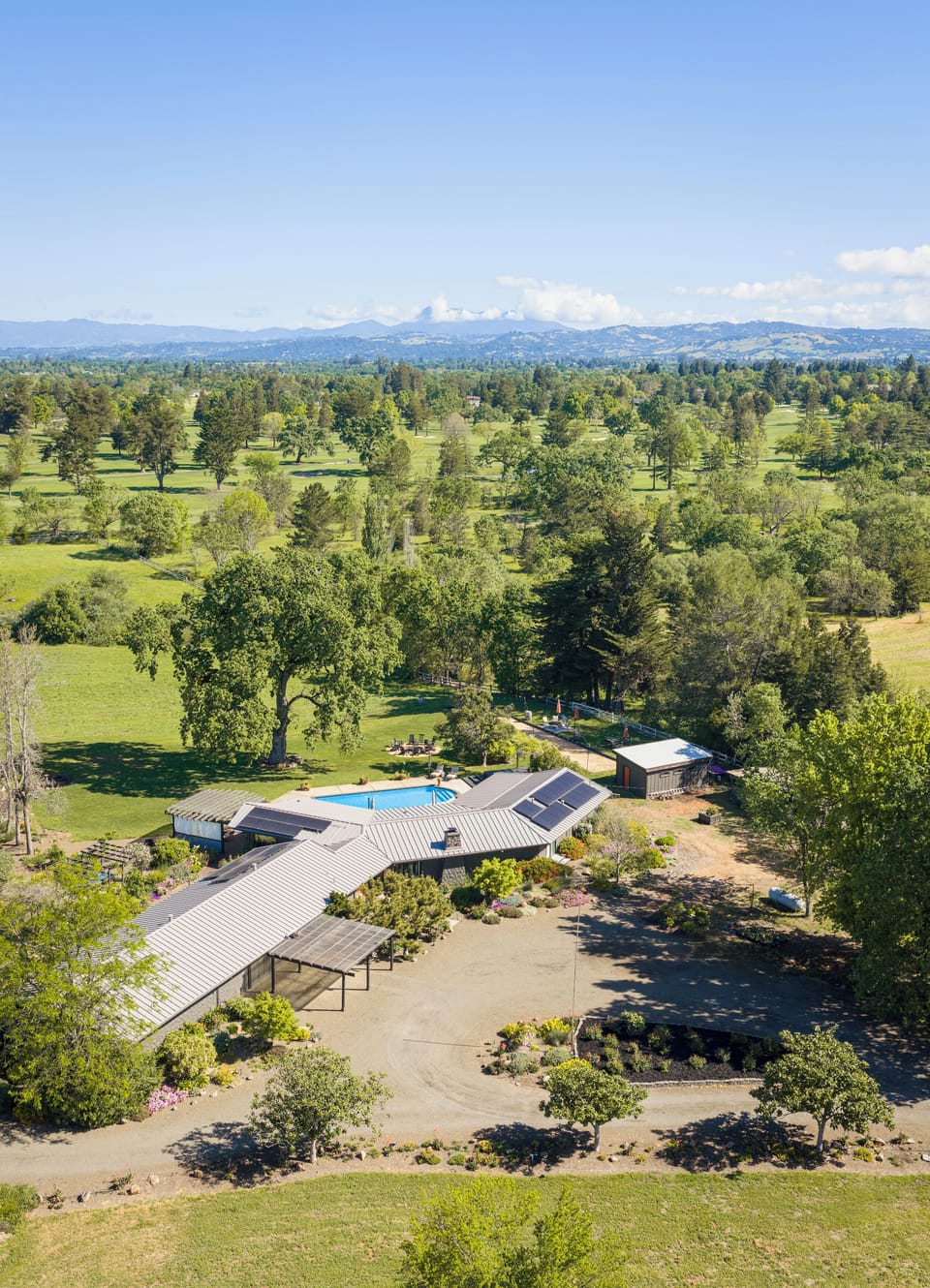Aerial view of a secluded ranch-style property nestled among towering evergreens and open meadows, with mountain ranges gracing the distant horizon under a clear blue sky.