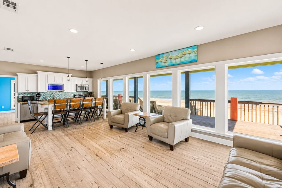 Open-concept living and dining area with large Gulf-facing windows, comfortable seating, wood-look floors and direct views of the beach and ocean.