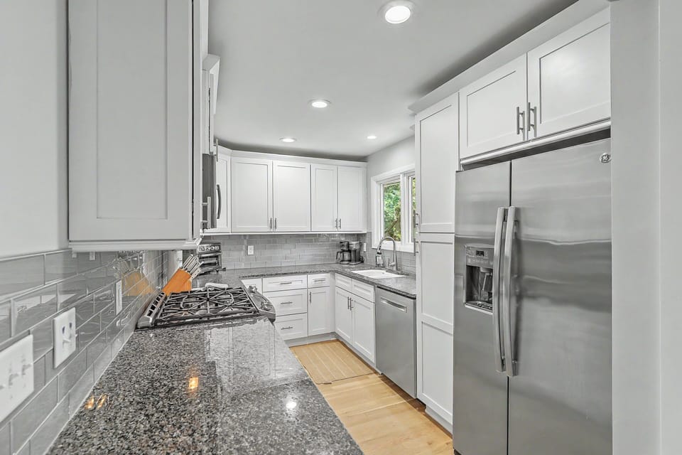 A full view of the fully remodeled galley kitchen highlights the long granite countertops, bright white cabinetry, a deep double sink, and sleek stainless steel appliances.