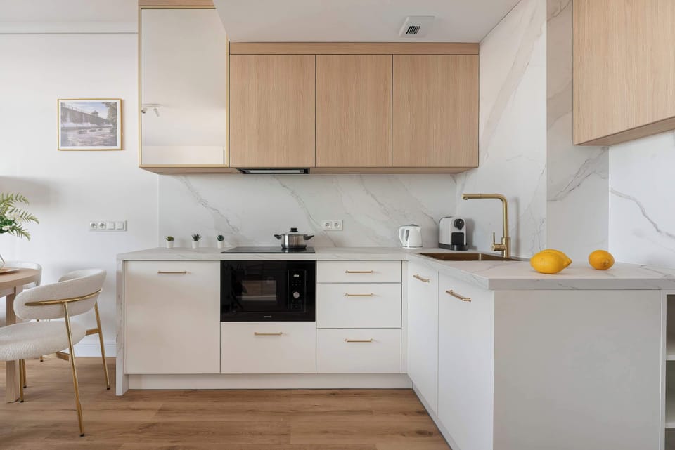 A sleek L-shaped kitchenette is designed with white cabinetry and light wood upper units. The marble-effect backsplash and gold-toned faucet add a touch of luxury to the functional space.