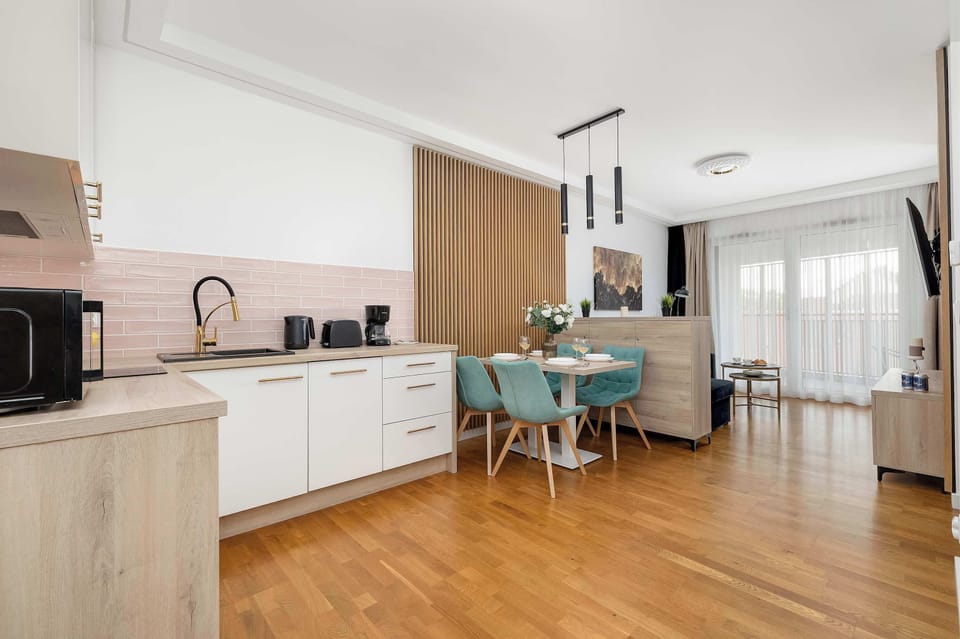 A bright, modern open kitchen with white cabinets and a wooden floor, featuring a central island with turquoise chairs. The space looks clean, spacious, and well-lit with natural light.