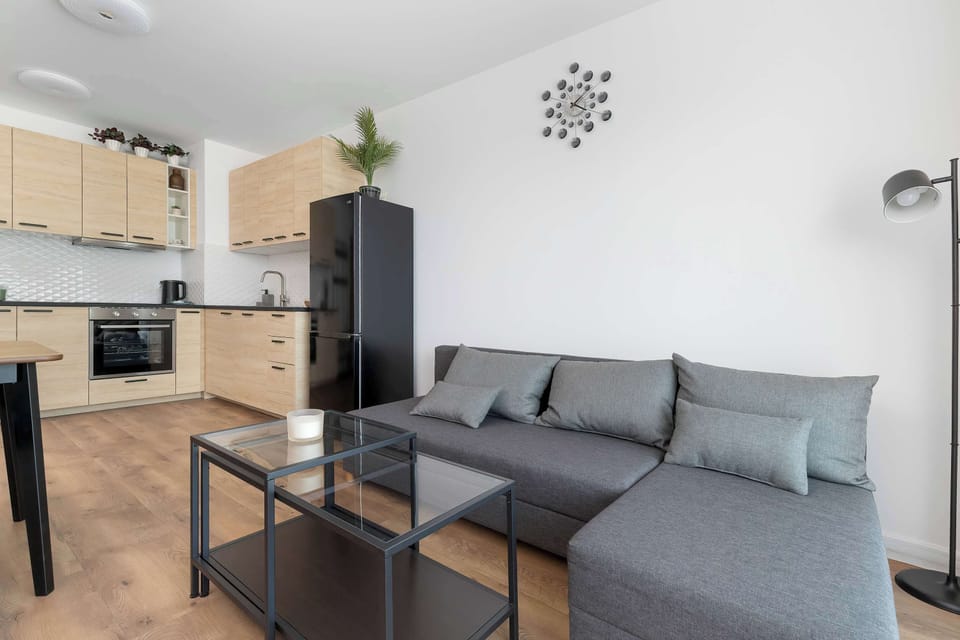 A spacious living room area taken from an angle that displays the gray corner sofa and kitchen background. The interior design relies on clean lines and neutral colors.